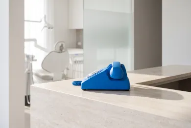 A blue desk phone on a travertine front desk, a dental chair behind glass